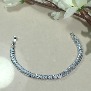 Pode incluir: Uma pulseira prateada com pedras preciosas retangulares e transparentes. A pulseira é curva e possui um fecho. As pedras preciosas estão próximas, criando uma faixa contínua e brilhante. A joia é exibida em uma superfície clara.