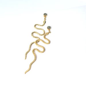 May include: A pair of gold-colored snake earrings. The earrings have a textured, hammered finish and a serpentine design. Each earring features a small, round, gray-green stone at the top. The earrings are displayed against a white background.