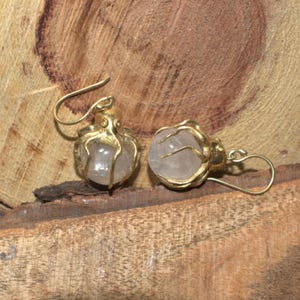 May include: A pair of gold-colored earrings featuring a clear, round stone. The earrings have a unique design, with the stone held in place by a decorative gold setting. The earrings are displayed on a wooden surface.