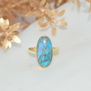 Könnte beinhalten: Goldener Ring mit einem ovalen türkisfarbenen Stein. Der Stein hat ein marmoriertes Aussehen mit Blau- und Brauntönen. Der Ring ist vor einem weißen Hintergrund mit goldenen floralen Akzenten platziert.
