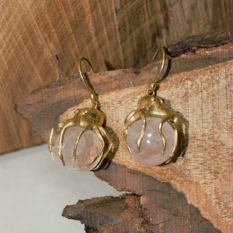 May include: A pair of gold-colored earrings featuring a round, pale pink gemstone. The gold setting has an intricate, organic design. The earrings are displayed against a textured, brown wooden surface.