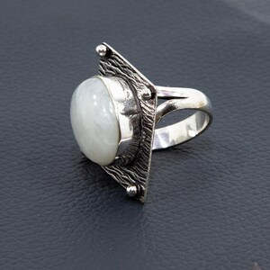 May include: A silver ring featuring a large, oval, milky white gemstone. The ring has a unique design with a textured, triangular accent on one side and a smooth band. The gemstone is set in a bezel setting.