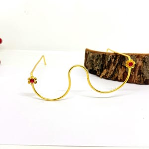 Könnte beinhalten: Goldfarbene Drahtbrillengläser mit einem einzigartigen, geschwungenen Design. Jedes Ende hat einen kleinen, roten Blumenakzent. Die Brille steht vor einem weißen Hintergrund mit einem Holzstück, das eine minimalistische Ästhetik zeigt.