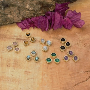 Könnte beinhalten: Eine Sammlung goldfarbener Ohrstecker mit verschiedenfarbigen Edelsteinen, darunter Rosa, Braun, Weiß, Blau, Grün, Schwarz und Lila. Die Ohrringe sind auf einer Holzoberfläche angeordnet, mit lila Blumen und einem Stück Rinde im Hintergrund.