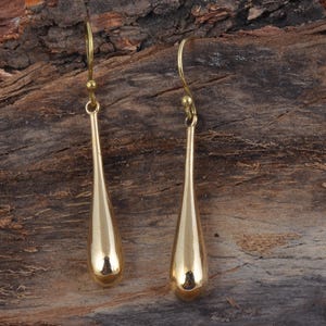 Könnte beinhalten: Ein Paar goldfarbene Tropfenohrringe. Die Ohrringe haben eine glatte, polierte Oberfläche und ein schlichtes, elegantes Design. Die Tropfenform ist länglich, mit einem abgerundeten Boden und einer schmalen Spitze. Die Ohrringe sind an einem goldfarbenen Haken befestigt.