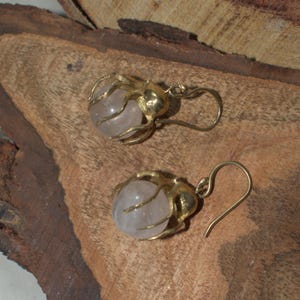 May include: A pair of gold-colored earrings with a light pink, spherical stone encased in a decorative wire frame. The earrings have a hook closure and are displayed on a wooden surface.