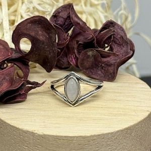 Könnte beinhalten: Ein silberner Ring mit einem hellfarbenen ovalen Stein in einem dekorativen Rahmen. Der Ring wird auf einer Holzoberfläche präsentiert, mit getrockneten burgunderfarbenen Blütenblättern im Hintergrund. Das Design des Rings ist schlicht und elegant.