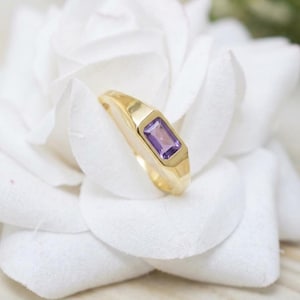 Könnte beinhalten: Ein goldener Ring mit einem einzelnen rechteckigen, im Smaragdschliff geschliffenen violetten Amethyst, der in eine glatte, polierte Metallschiene gefasst ist. Der Ring ist mittig platziert und zeigt den leuchtenden Farbton des Steins.