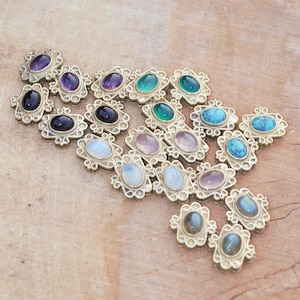 Könnte beinhalten: Eine Sammlung verzierter, goldfarbener Schmuckkomponenten mit verschiedenen farbigen Edelsteinen. Die Edelsteine umfassen Amethyst, Onyx, Türkis und Mondstein, die in dekorativen Rahmen gefasst sind. Die Komponenten sind auf einer hellbraunen Holzoberfläche angeordnet.