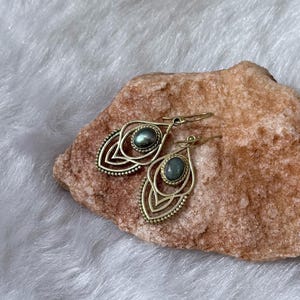 May include: A pair of gold-colored earrings with a teardrop design. Each earring features a central oval-shaped, gray-green stone. The earrings are placed on a rough, reddish-brown rock against a white, fluffy background.