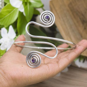May include: A silver-toned metal arm cuff featuring a continuous wire design that forms two prominent spirals at each end. Each spiral is centered with a small, round, deep purple amethyst gemstone, creating a symmetrical and polished appearance.
