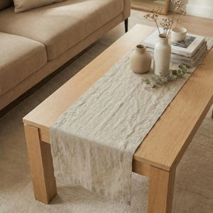 Könnte beinhalten: Ein hellbeige Leinen-Tischläufer auf einem hellen Holz-Couchtisch. Der Tisch ist mit einer kleinen beigen Vase, einer weißen Vase und Büchern dekoriert. Im Hintergrund befindet sich ein beiges Sofa.
