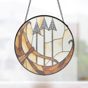 May include: A round stained glass window with a black metal frame and chain for hanging. The design features a Viking ship in shades of brown and tan, with three gray arrows against a white and cream background.