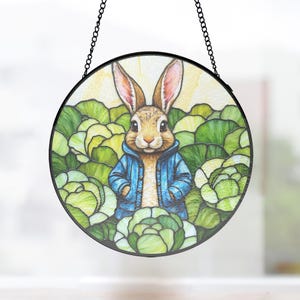 Può includere: Un pannello rotondo in vetro colorato raffigurante un coniglio con una giacca blu circondato da cavoli verdi. Il coniglio ha il pelo marrone e lunghe orecchie. Il pannello è incorniciato in nero e appeso a una catena nera.