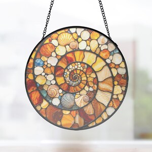 May include: A circular stained glass suncatcher featuring a spiral pattern composed of various amber, orange, yellow, and white glass pieces shaped like seashells and pebbles, all held together by dark metal cames and suspended by a black chain.
