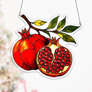 Op de afbeelding: Gebrandschilderd glasraam met twee granaatappels, één heel en één gehalveerd, met rode zaden. Het ontwerp omvat een bruine tak met groene en gele bladeren, opgehangen aan een ketting. De granaatappels zijn helderrood.