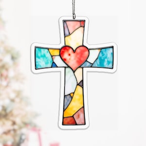 Pode incluir: Um enfeite de cruz de vitral com um coração no centro. A cruz apresenta uma borda branca e é feita de peças de vidro coloridas em tons de vermelho, azul, amarelo e verde. Uma corrente prateada é anexada para pendurar.