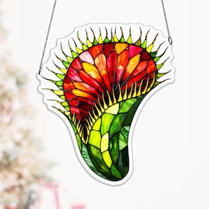 May include: Stained glass artwork of a stylized flower. The centre is red, orange, and yellow, with green and yellow accents. The piece is outlined in white and suspended by a chain.