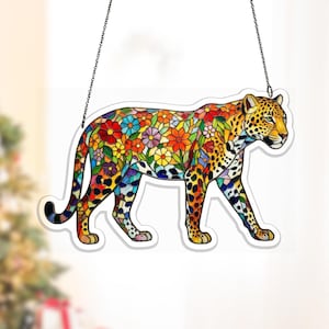 Puede incluir: Decoración de leopardo de estilo vidriera. El cuerpo del leopardo está lleno de un vibrante estampado floral en rojo, amarillo, azul y morado. El leopardo está delineado en negro y cuelga de una cadena.