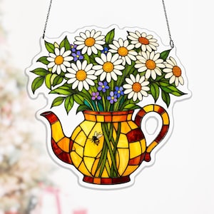 Può includere: Vetrata artistica raffigurante una teiera gialla piena di margherite bianche, fiori blu e foglie verdi. La teiera presenta un motivo geometrico nei toni del giallo, arancione e rosso. Una piccola ape è visibile sulla teiera. L'opera d'arte è appesa a una catena.