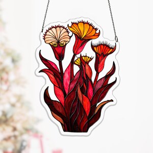 May include: A stained glass window hanging featuring a floral design. The artwork showcases vibrant red, orange, and yellow flowers with detailed leaves. The piece is outlined in white and suspended by a delicate chain, perfect for home decor.