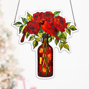 Peut inclure: Vitrail représentant un bouquet de roses rouges et deux cardinaux rouges dans une bouteille rouge. Les feuilles sont vertes et jaunes. L'art de la fenêtre est suspendu à une chaîne.