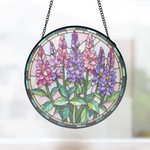 Può includere: Un acchiappasole circolare in vetro colorato raffigurante un grappolo di fiori di lupino rosa e viola con foglie verdi. Il disegno è racchiuso in una cornice di metallo scuro con bordo decorativo e appeso a una catena di metallo nera.