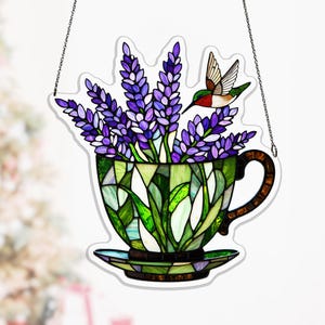 May include: Stained glass teacup suncatcher with lavender and a hummingbird. The teacup is green and white with a brown handle and saucer. The lavender is purple. The hummingbird has red, green, and brown feathers. A chain is attached for hanging.