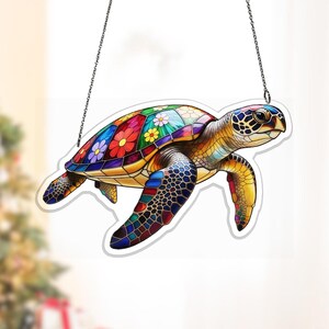 May include: A colorful sea turtle decoration with a stained glass effect. The turtle's shell is adorned with vibrant flowers in red, yellow, blue, and purple. The turtle is suspended by a silver chain, perfect for hanging. The design features a variety of colors and patterns.