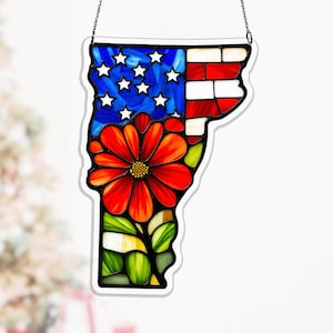Può includere: Suncatcher in vetro colorato a forma di Vermont, con la bandiera americana e un fiore rosso. Il suncatcher è bordato di bianco e appeso a una catena d'argento. Il fiore ha un centro giallo e foglie verdi.