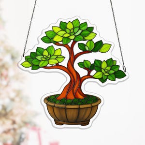 May include: A stained glass bonsai tree suncatcher. The tree has a brown trunk and branches with green leaves. It is planted in a brown pot filled with green pebbles. The suncatcher hangs from a silver chain.