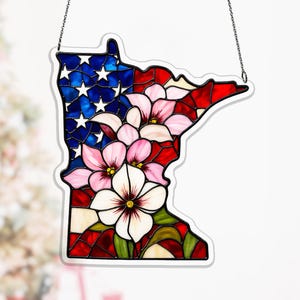May include: Stained glass art piece in the shape of the state of Minnesota, featuring a design of the American flag and pink and white flowers. The piece is outlined in black and hangs from a chain.