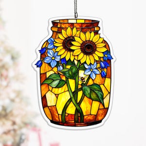 Puede incluir: Un diseño de tarro de cristal manchado con girasoles, flores azules y una abeja. El tarro está lleno de vidrieras amarillas y naranjas, con una cadena para colgar. Los girasoles tienen pétalos amarillos y centros marrones.