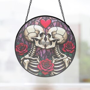 Op de afbeelding: Ronde decoratie met zwarte lijst, met twee kussende skeletten en een rood hart erboven. Rode rozen en een paarse achtergrond met glas-in-lood effect. Opgehangen aan een zwarte ketting.