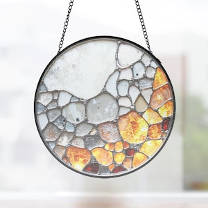 May include: Round stained glass window hanging with a black metal frame and chain. The design features a mosaic of stones in shades of white, gray, orange, and red. The glass has a textured appearance, creating a unique decorative piece.