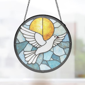 Puede incluir: Vidriera redonda que representa una paloma blanca en vuelo sobre un sol amarillo y un fondo azul. La ventana está enmarcada en negro y cuelga de una cadena. El diseño recuerda a una vidriera de iglesia.
