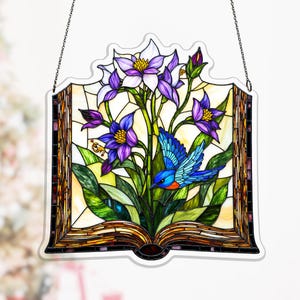May include: Stained glass panel featuring a blue bird, purple flowers, and green leaves. The artwork is shaped like an open book with a decorative border and hanging chain. The panel has a colorful, mosaic-like appearance.