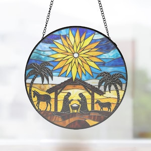 May include: Round stained glass window art depicting a nativity scene. The artwork features a bright yellow star, palm trees, and silhouettes of figures in a stable. The piece is framed in black and hangs from a chain.