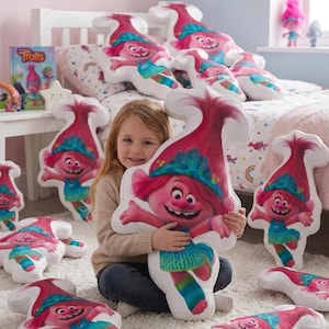 May include: A young girl smiles while holding a large Poppy pillow from the Trolls movie. Several other Poppy pillows are scattered around, along with a Trolls book and figurines. The pillows feature Poppy's pink hair, blue dress, and smiling face.