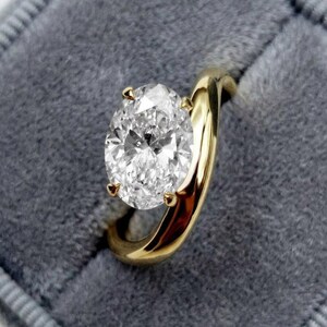 Può includere: Un anello di fidanzamento in oro con un grande diamante taglio ovale. L'anello è incastonato in una classica montatura a quattro griffe e poggia su una superficie di velluto grigio. La fascia è in oro lucido.