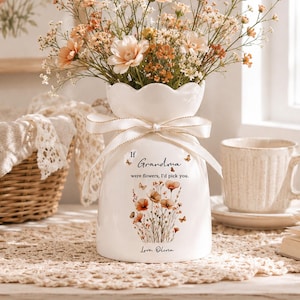 Könnte beinhalten: Eine weiße Keramikvase mit gewelltem Rand und einem cremefarbenen Band um den Hals. Die Vase zeigt eine Blumenillustration und den Text: If Grandma were flowers, I'd pick you. Love, Olivia. Sie ist mit verschiedenen Wildblumen gefüllt.