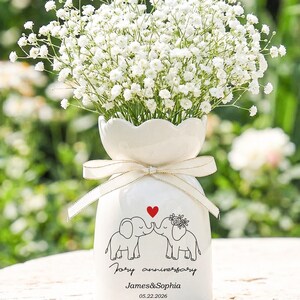 May include: A white ceramic vase with a scalloped rim and a cream-colored ribbon tied around the neck. The vase features a black line drawing of two elephants with a red heart, the text "Jory anniversary", "James&Sophia", and "05.22.2026".