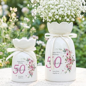 May include: Two white ceramic vases with floral arrangements. The vases are decorated with pink text that reads "Happy 50th Birthday" and "Olivia". A floral design and a ribbon add to the decorative look.