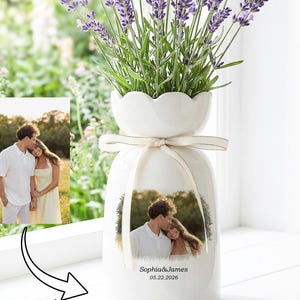 May include: A white ceramic vase with a scalloped rim, filled with purple lavender stems. The vase features a custom printed photograph of a couple, a cream-colored ribbon tied around the neck, and the text Sophia & James 05.22.2026 printed below.