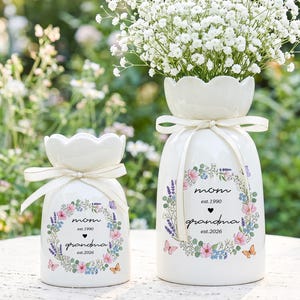Op de afbeelding: Twee witte keramische vazen, een grote en een kleine, met bloemmotieven en de woorden "mama" en "oma". De grotere vaas bevat witte bloemen. Beide vazen hebben een lint om de hals geknoopt.