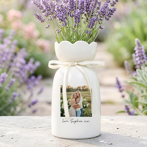 Op de afbeelding: Een witte keramische vaas met een geschulpte bovenkant, vastgebonden met een crèmekleurig lint en een gedrukte foto. De vaas bevat paarse lavendel. De tekst "Love Sophia xx" is eronder gedrukt.