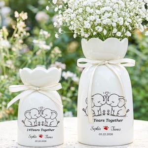 Könnte beinhalten: Zwei weiße Keramikvasen mit Blumendesign und Schleifenakzenten. Die größere Vase enthält weiße Blumen. Jede Vase zeigt Elefanten-Cartoons und den Text "Years Together" und "14 Years Together".