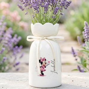 May include: A white ceramic vase with a scalloped rim and a cream-colored ribbon tied around the neck. The vase features a graphic of Minnie Mouse blowing a kiss and the name Isabella printed in black script on the front, filled with lavender.