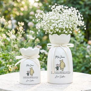 Op de afbeelding: Twee witte keramische vazen met een Winnie de Poeh-ontwerp en tekst. De grotere vaas bevat witte bloemen. Beide vazen hebben een geschulpte bovenkant en zijn vastgebonden met een lint. De tekst bevat de naam "Mrs. Jones" en een citaat van Winnie de Poeh.