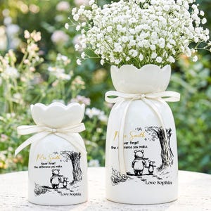 Può includere: Due vasi in ceramica bianca con un motivo floreale e un nastro legato attorno al collo. I vasi presentano un'illustrazione di Winnie the Pooh e il testo "Mr. Smith, Non dimenticare mai la differenza che fai, Love Sophia". Un vaso è pieno di fiori bianchi.